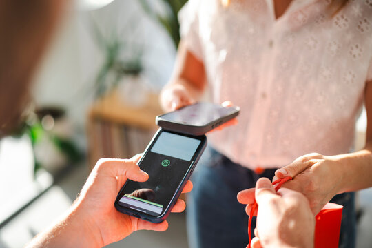 A customer completes a contactless transaction by tapping a smartphone on a mobile payment terminal held by a retail worker. A shopping bag is visible in hand.