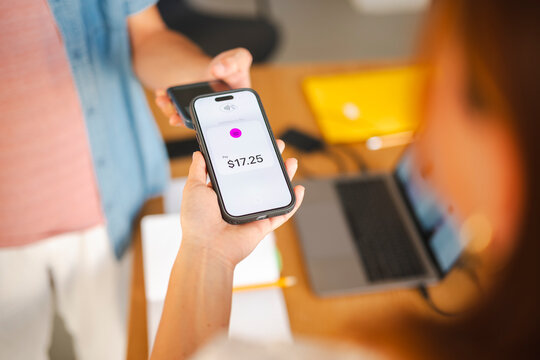 A customer uses their smartphone to make a secure contactless digital payment for their purchase at a local shop. A merchant holds the terminal for the transaction.