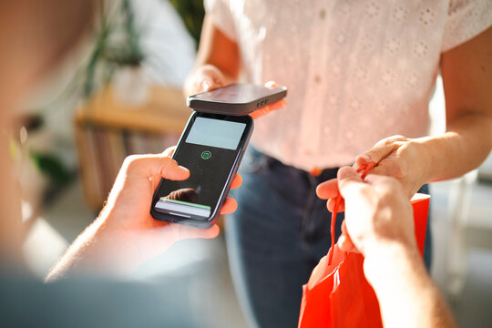 A store clerk holds a mobile payment terminal for a customer to pay with their phone. The customer receives a red shopping bag after completing the purchase.