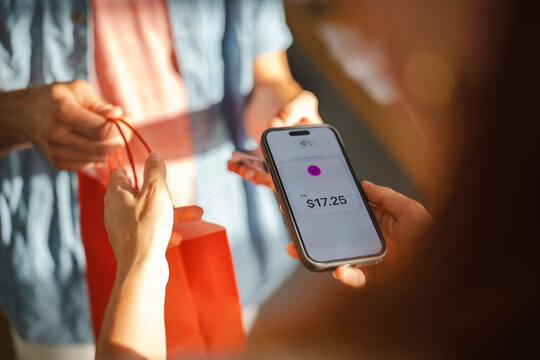 A customer pays for a purchase using a mobile device for a contactless transaction. The shop assistant hands over a red shopping bag at the retail checkout counter.