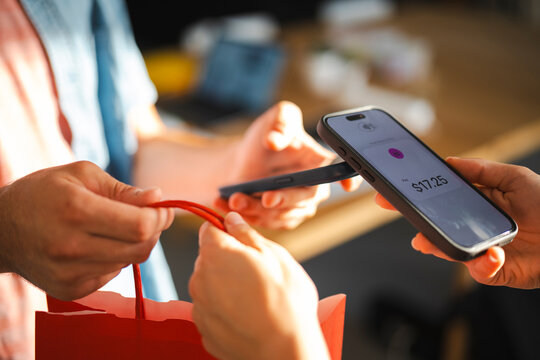 A customer uses a smartphone for a contactless payment at a store. The cashier hands over a red shopping bag as the transaction completes in the retail shop.
