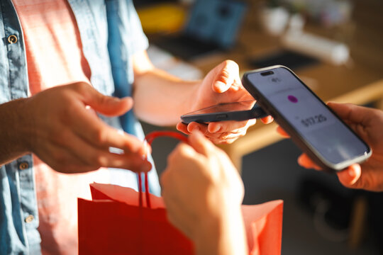 A customer uses a smartphone to complete a contactless payment for a purchase at a store. The merchant holds a red shopping bag during the digital transaction process.