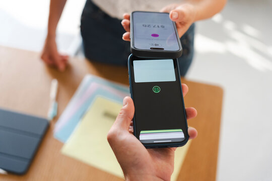 A customer completes a secure contactless payment by tapping their smartphone against a merchant's mobile device. This simple transaction occurs at a wooden workspace.