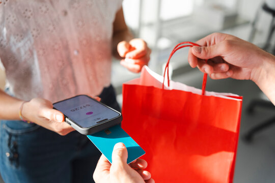A retail worker hands a red shopping bag to a customer. The customer is making a payment by tapping their credit card onto a mobile phone used as a point of sale.