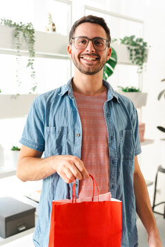 A happy young man with glasses smiles while holding a red shopping bag in a bright, plant-filled indoor setting. He is enjoying his recent purchase in the store.