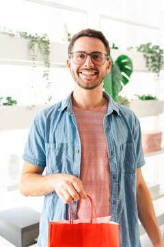 A young man with glasses smiles while holding a red shopping bag in a bright, modern shop. He is wearing a blue denim shirt and feels happy with his recent purchase.