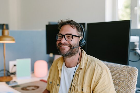 Man wearing a headset and hearing aid, smiling while working at a desk with blank monitors and copy space for customer service and holiday campaigns
