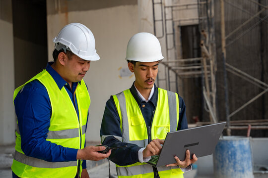 Two Asian civil engineers in safety helmets and reflective vests using a laptop for project planning and discussing at a construction site