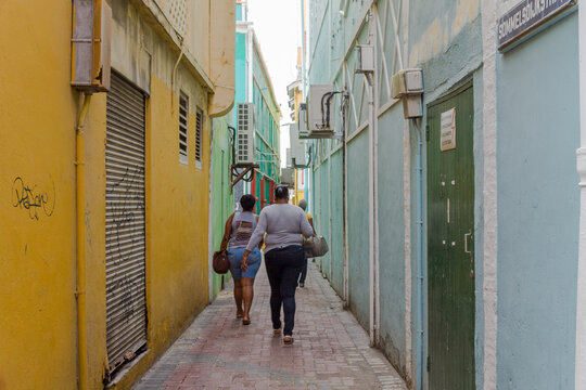 Willemstad, Curacao, Curacao - 06 November 2014: View of a narrow alleyway in Willemstad with vibrant yellow and light blue walls, brick paving, and people walking.