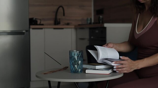 A woman sits calmly in a cozy kitchen, holding a book with a serene and focused mood. The side view highlights the wooden table, ceramic cup, and paper book, creating an educational and hom.