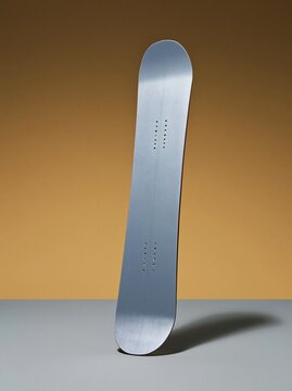 Plain snowboard standing upright with a high-gloss finish and blank topsheet, set against a golden orange background for winter retail mockup and copy space