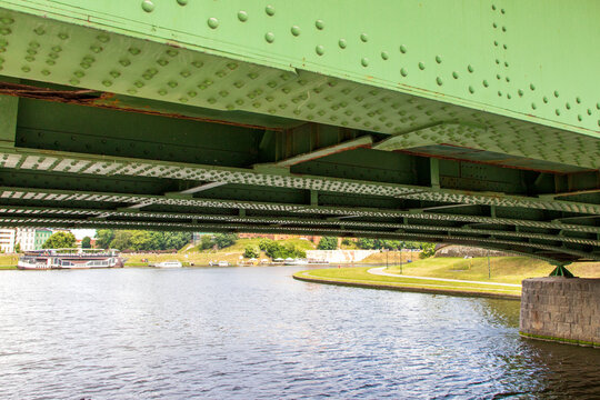 A close-up view of a large, painted green steel plate bridge seen from a boat passing beneath it. with rows of structural rivets. A view from astern to the churned-up water and the green riverbanks. 
