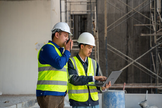 Two Asian construction engineers coordinating on site, one speaking on a walkie-talkie while the colleague uses a laptop for project management.