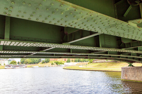 A close-up view of a large, painted green steel plate bridge seen from a boat passing beneath it. with rows of structural rivets. A view from astern to the churned-up water and the green riverbanks. 