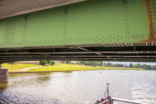A close-up view of a large, painted green steel plate bridge seen from a boat passing beneath it. with rows of structural rivets. A view from astern to the churned-up water and the green riverbanks.  