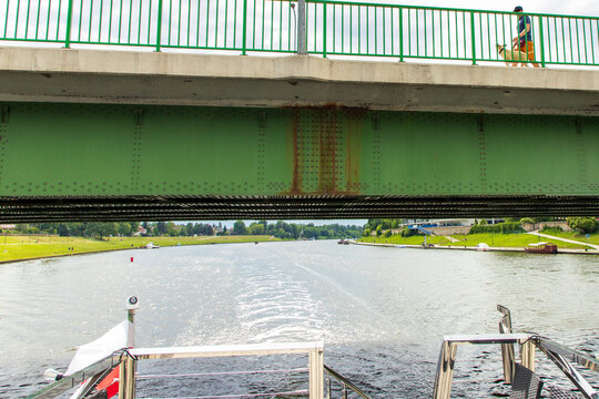 A green-and-gray bridge seen from a boat passing beneath it. A pedestrian with a dog is walking along the green railing. A view from astern to the churned-up water and the green riverbanks. 