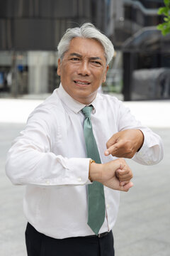 Angry and upset senior asian businessman worried about time while pointing at his wristwatch in an urban central business area setting, concept image for punctuality, coming late