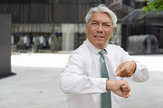 Angry and upset senior asian businessman worried about time while pointing at his wristwatch in an urban central business area setting, concept image for punctuality, coming late