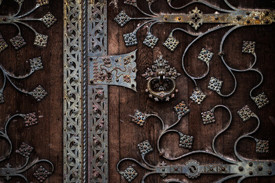 Metal design across an antique wooden door. Metal design with ornamental wrought iron scrollwork. Historic door metal design with decorative studs. Gothic entrance with metal design and symmetry.