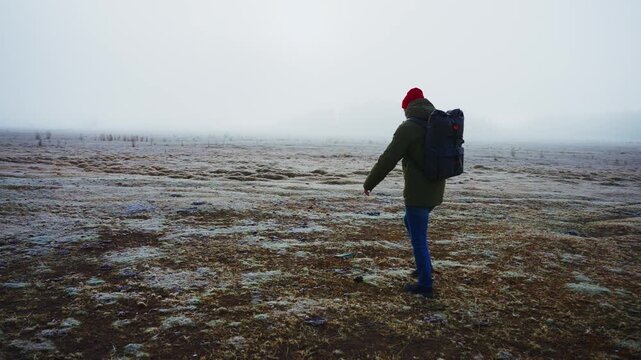 Lone hiker walks across a frozen, misty field. They wear a red cap, green jacket, and blue jeans. Backpack suggests outdoor adventure or travel. Atmosphere feels quiet, cold, and slightly eerie