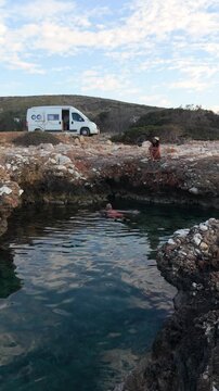 In the beautiful surroundings of Paros Island, adventurous swimmers enjoy the crystal-clear waters of natural pools. A couple relaxes by the ocean with a camper van motorhome in the background