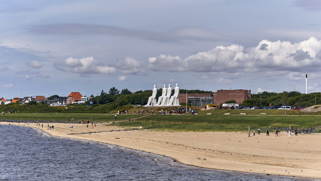 Esbjerg, Denmark - 28 July 2026: View of the Mennesket ved Havet sculpture by Svend Wiig Hansen, featuring four large white figures on the coast with people walking nearby under a cloudy sky.