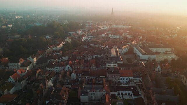 Warm sunrise over Bruges, Belgium, reveals dense red rooftops, historic houses and distant church spires across the atmospheric medieval cityscape at dawn.