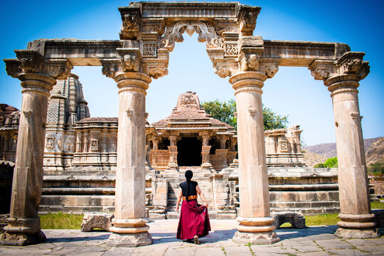 lady in a long skirt with long braided hair standing with her back four intricately carved stone pillars at Sahastrabahu Temple, Udaipur, Rajasthan