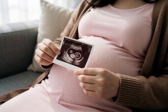 Close up of pregnant woman holding ultrasound scan photo in hands. Expectant mother looks at baby sonogram sitting on couch. Happy female shows fetal development result during pregnancy. Motherhood