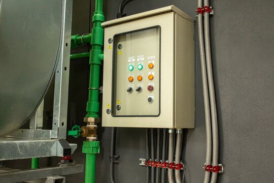 Industrial electrical control panel with buttons and indicator lights mounted on wall near pipes, automation equipment concept