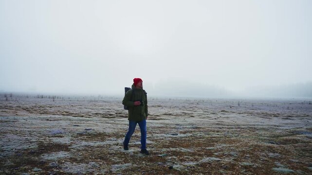 Person walks through foggy, frost-covered field, wearing red hat and green jacket. They adjust hat, step forward with backpack, moving slowly through misty landscape. Ground covered with frost