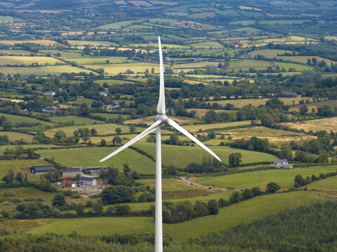 Aerial view of a wind turbine standing in the green countryside with rolling hills, agricultural fields, and scattered farm buildings in Cavan, County Cavan, Ireland.