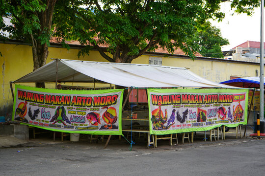 Traditional Indonesian street food tent stall, known as warung makan pecel lele, featuring colorful hand-painted banners depicting menu items like chicken, catfish, and fish on a sunny urban roadside.