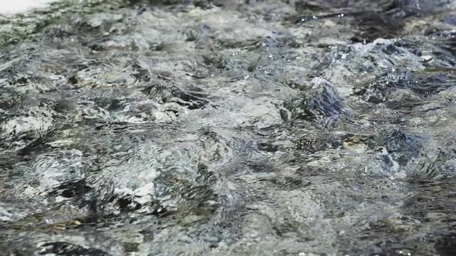 Water flows over rough stones. Tiny bubbles form as current moves fast. Surface reflects light in shimmering patterns. Ideal for nature, adventure, or eco-themed projects. Can be used in travel