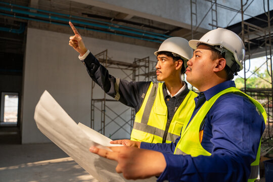 Two Asian construction engineers in safety helmets and reflective vests holding a large blueprint and pointing upwards while inspecting the building site.