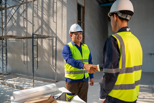 Two smiling Asian construction engineers shaking hands in agreement at a building site, with blueprints and a tablet on a table.