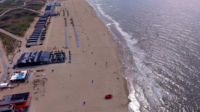 High angle horizontal aerial drone video clip of beach pavilion, rescue station, boat, beach cabins, North Sea waves, and sand dunes in Katwijk aan Zee, South Holland, Netherlands, West Europe