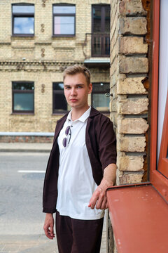 Young blonde man in brown clothing standing European city street with classic architecture and urban lifestyle atmosphere.
