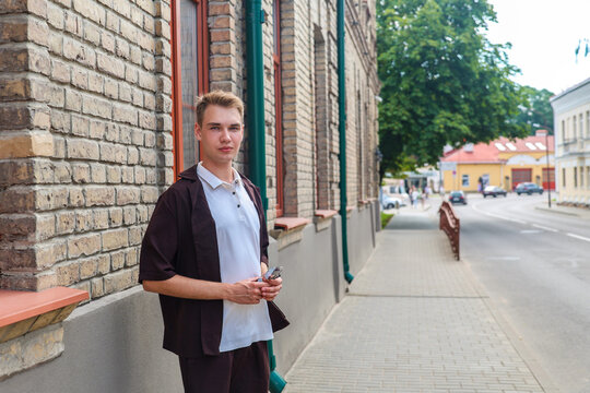 Young blonde man brown clothing standing on a European city street with classic architecture and urban lifestyle atmosphere.