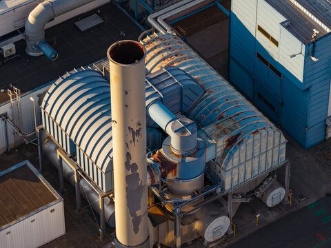 Aerial view of industrial infrastructure featuring a tall exhaust stack and large ventilation units at a manufacturing facility in Genk, Flanders, Belgium.