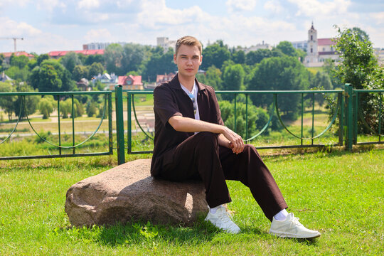 Handsome 20-year-old young man sitting large stone in a beautiful green park outdoors relaxing and enjoying peaceful nature scenery on sunny day.