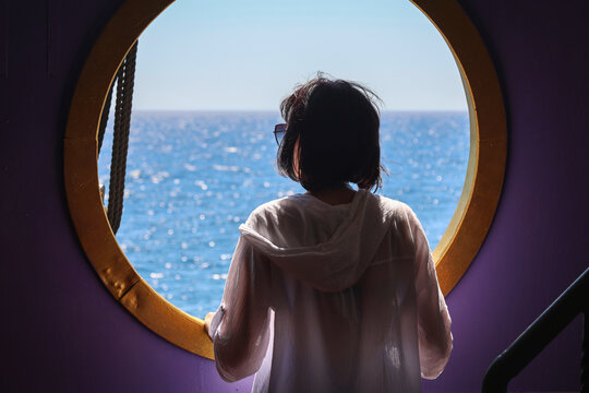Beautiful woman standing next to a large open round porthole window on a cruise ship deck enjoying ocean breeze and travel vacation.