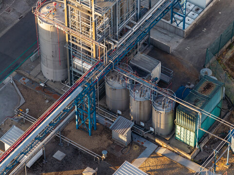 Aerial view of industrial infrastructure featuring stainless steel storage tanks, complex piping systems, and metal structures at a manufacturing facility in Genk, Flanders, Belgium.