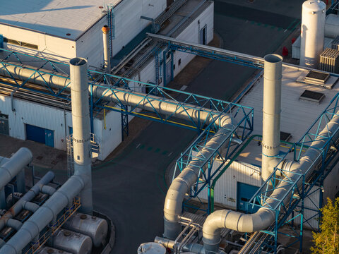 Aerial view of industrial infrastructure featuring complex piping systems, metal chimneys, and manufacturing buildings in Genk, Flanders, Belgium.