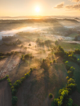 Aerial view of the misty landscape at sunrise, with golden sun rays illuminating the rolling hills, lush green trees, and agricultural fields in Loubressac, Occitanie, France.