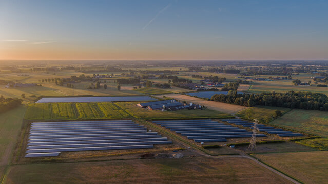 Aerial view of a solar farm with rows of photovoltaic panels situated in a rural landscape with green fields and farm buildings under the soft light of the golden hour in the Netherlands.