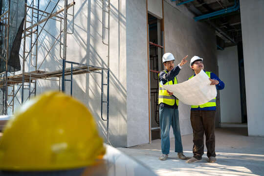 Two Asian construction engineers discussing a blueprint and pointing at the building site, with a blurred yellow hard hat in the foreground.