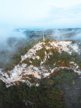 Aerial view of Roc Coulon Point de vue with its communication tower atop a rocky cliff surrounded by dense forest and low-hanging clouds in Saint-Sozy, Occitanie, France.