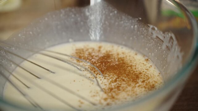 A close up macro shot shows ground cinnamon falling in a fine stream into creamy white pastry cream in a transparent glass mixing bowl.