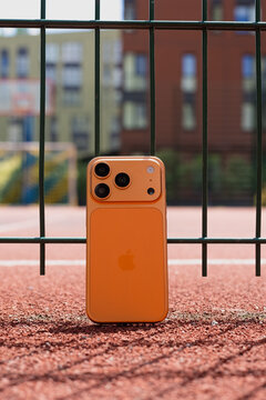 KYIV, UKRAINE - July 10, 2026 : Apple iPhone 17 Pro in Cosmic Orange color leaning against a green wire fence on a red running track, outdoor sport concept.